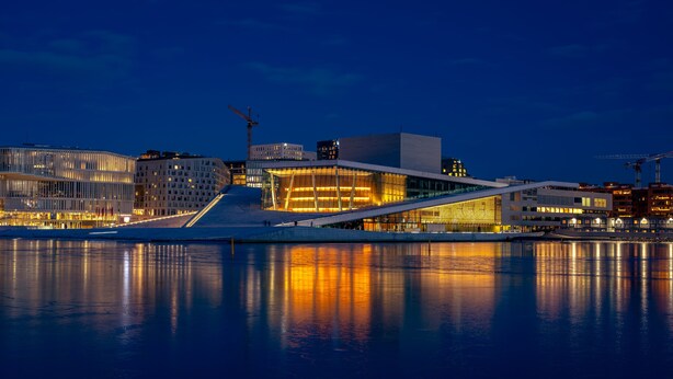 A modern waterfront building with a glass facade reflecting lights, a waterfront promenade, and adjacent urban buildings. - Tjuvholmen II As Home (Oslo)