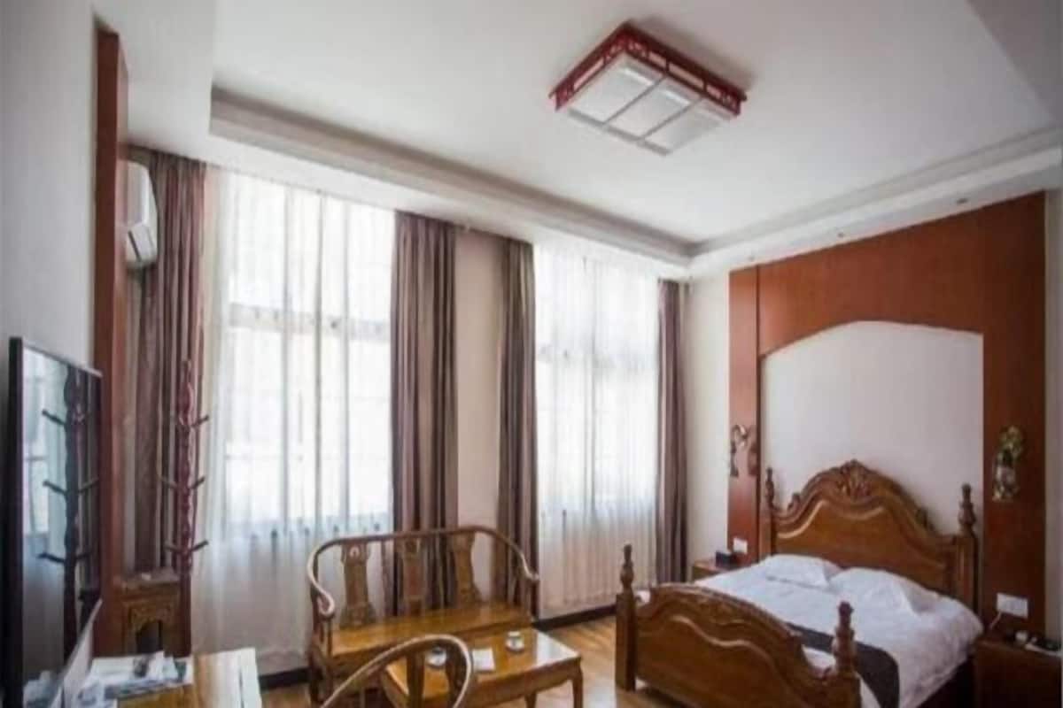 A hotel room with a wooden bed, a wooden table, a chair, a TV, and large windows with curtains.