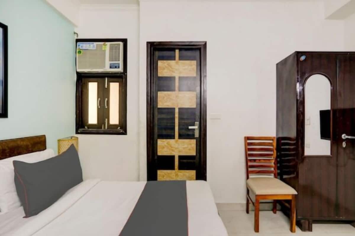 A hotel room with a bed, a chair, a wardrobe, and a window with an air conditioning unit.