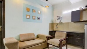 A compact living space with a sofa and armchair, a kitchenette with cabinets, and a wall-mounted shelf. - Victoria Palace (New Delhi)