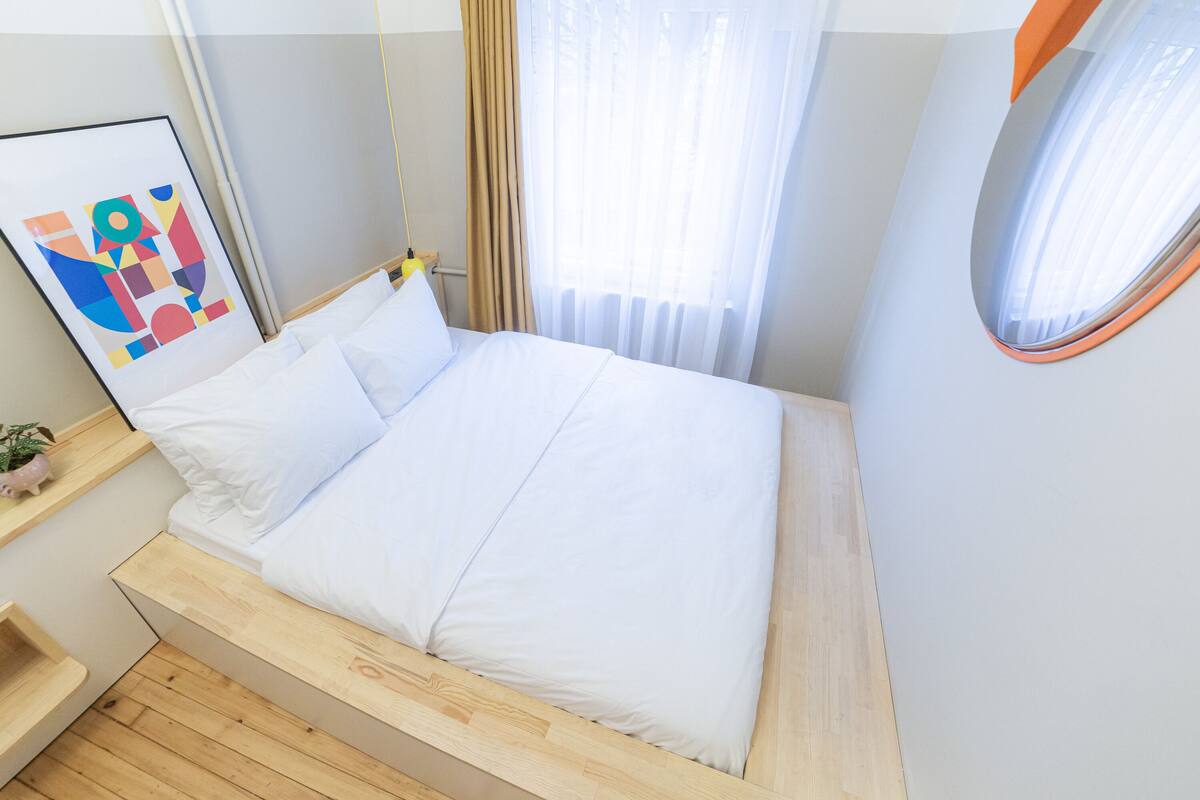 A modern bedroom with a wooden bed frame, white bedding, a small bedside table with a plant, and a colorful abstract painting on the wall.