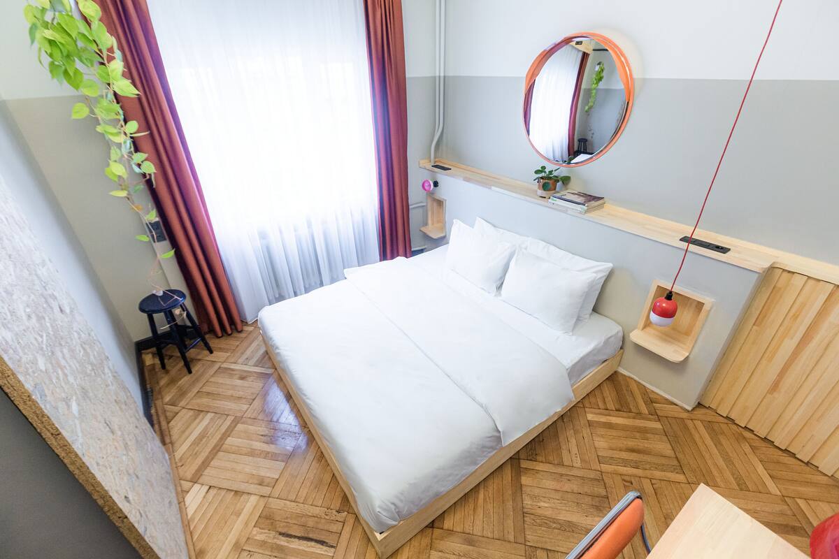 A modern hotel room with a wooden herringbone floor, a large bed with white linens, a round mirror on the wall, and a small shelf with a plant and a red hanging light.