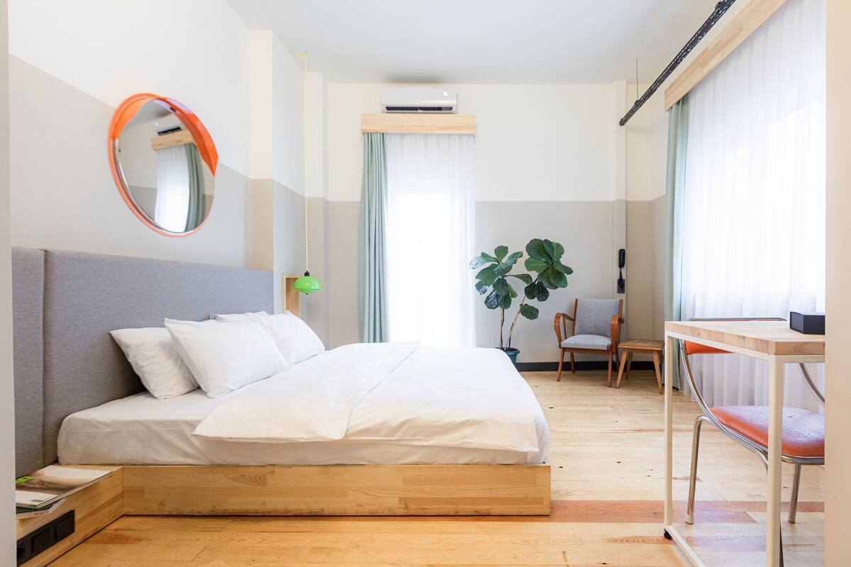 A modern hotel room with a bed, a desk, a chair, a large plant, and a mirror.