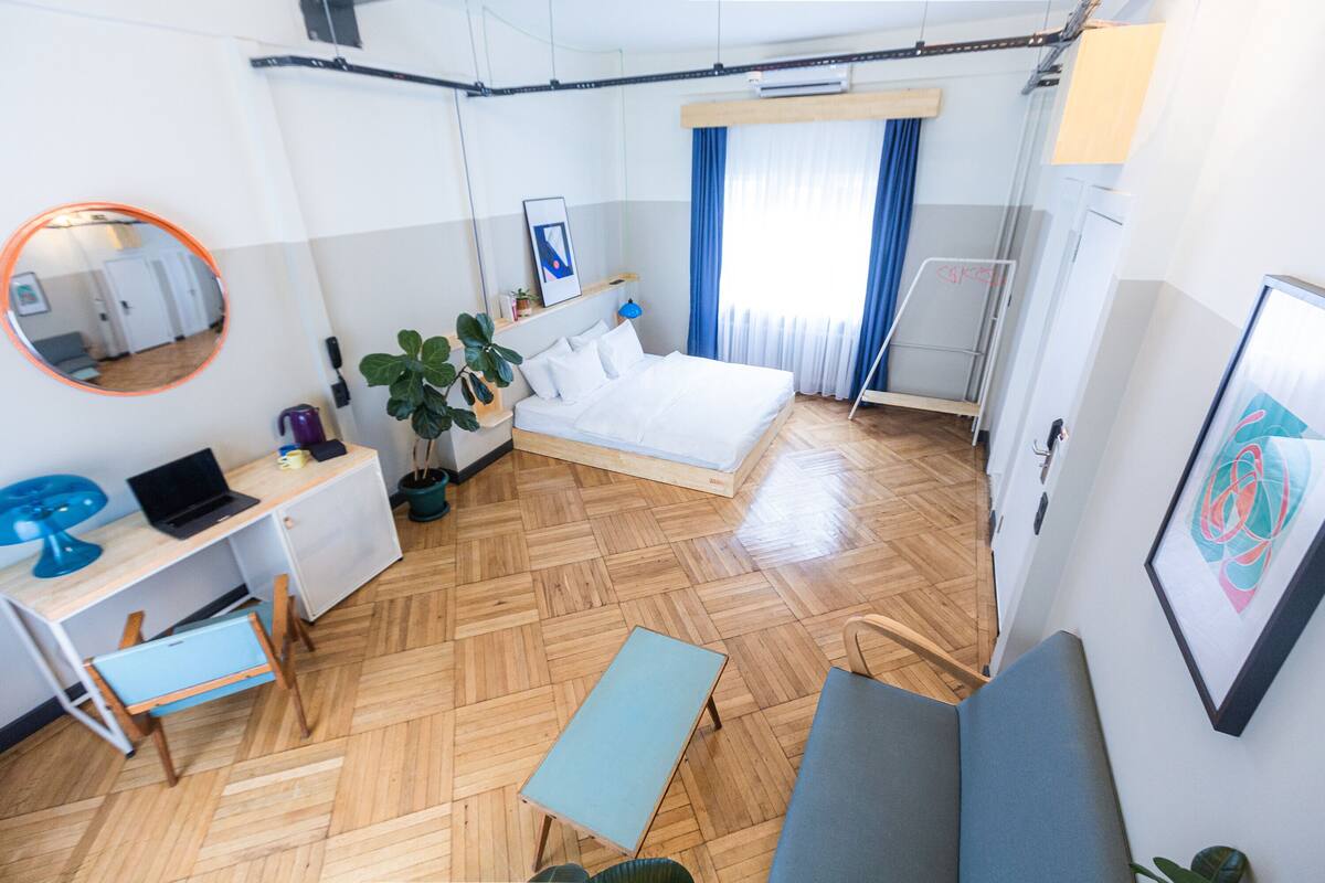 A compact hotel room with a bed, a desk with a laptop, a small table, a sofa, a mirror, and a potted plant.