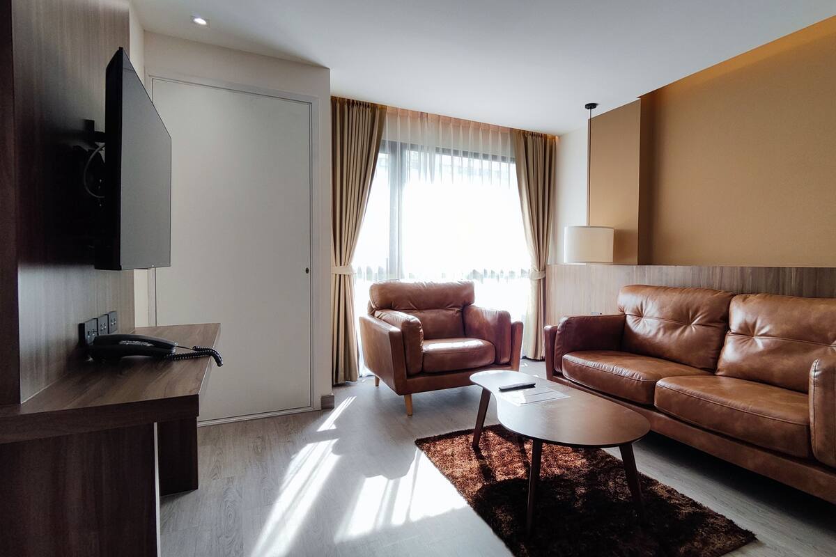 A modern hotel room with a brown leather sofa, a smaller armchair, a coffee table, a flat-screen TV mounted on the wall, and a large window with curtains.