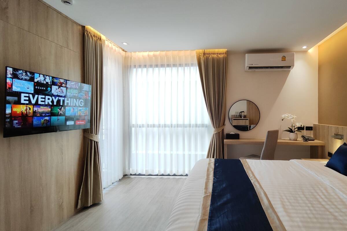 A hotel room with a large bed, a desk with a chair, a TV mounted on the wall, and an air conditioning unit.