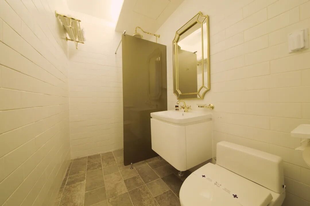 A bathroom with white tiled walls, a white sink with gold fixtures, a gold-framed mirror, and a toilet with a gold emblem.