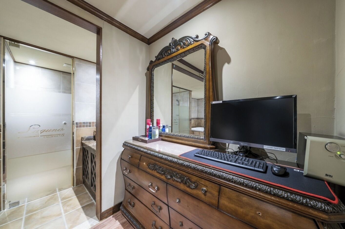 A bathroom with a wooden vanity, a large mirror, a flat-screen TV, and toiletries on the counter.