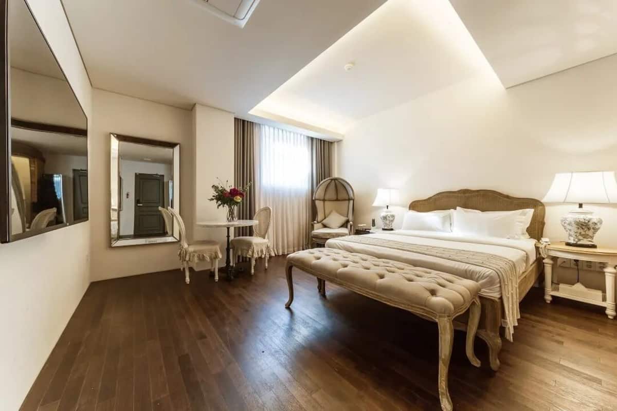 Una camera d'albergo con un letto grande, un'area salotto con una sedia e un tavolo, uno specchio e una finestra con tende.