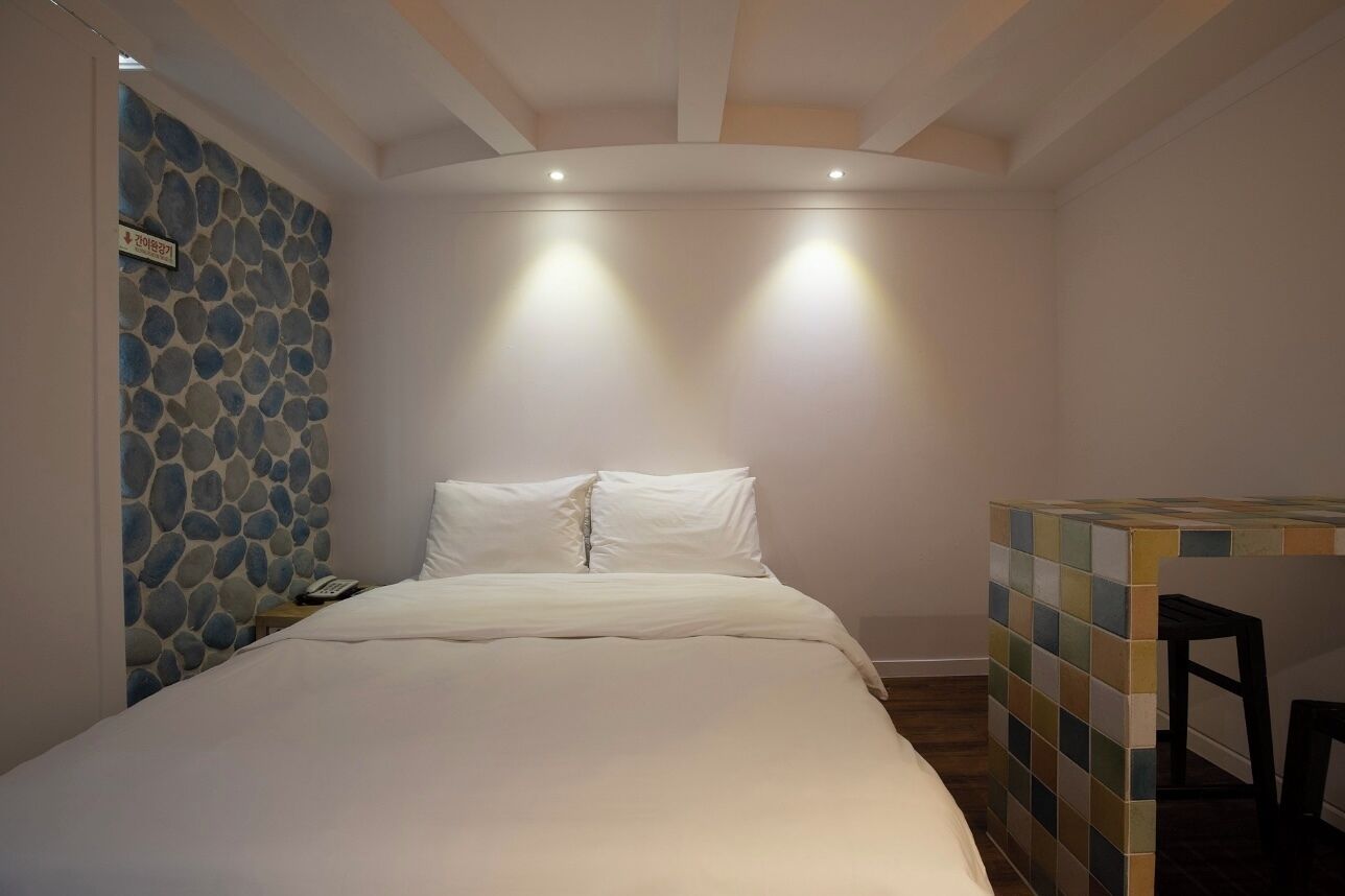 A bedroom with a bed, a nightstand, a wall with a stone pattern, and a small table with a chair.