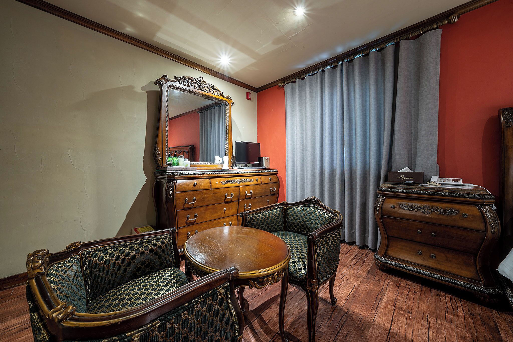 A room with a wooden dresser, two upholstered chairs, a round wooden table, and a large mirror.