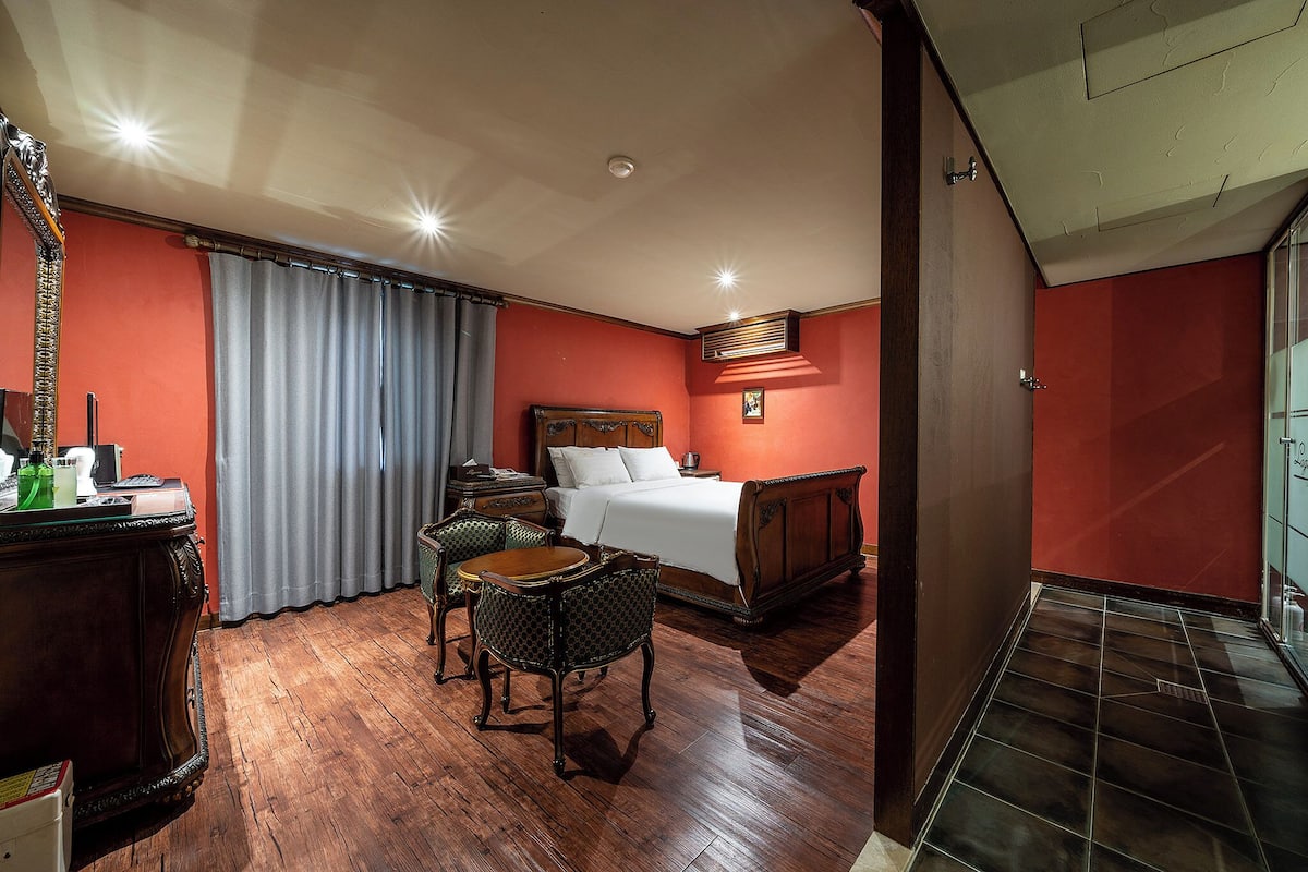 Una camera d'albergo con pavimento in legno, un letto con lenzuola bianche, una scrivania con uno specchio e una sedia.