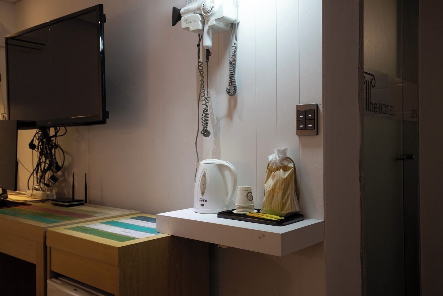 A hotel room with a flat-screen TV mounted on the wall, a hairdryer, a kettle, and a tray with a cup and a bag of bread.