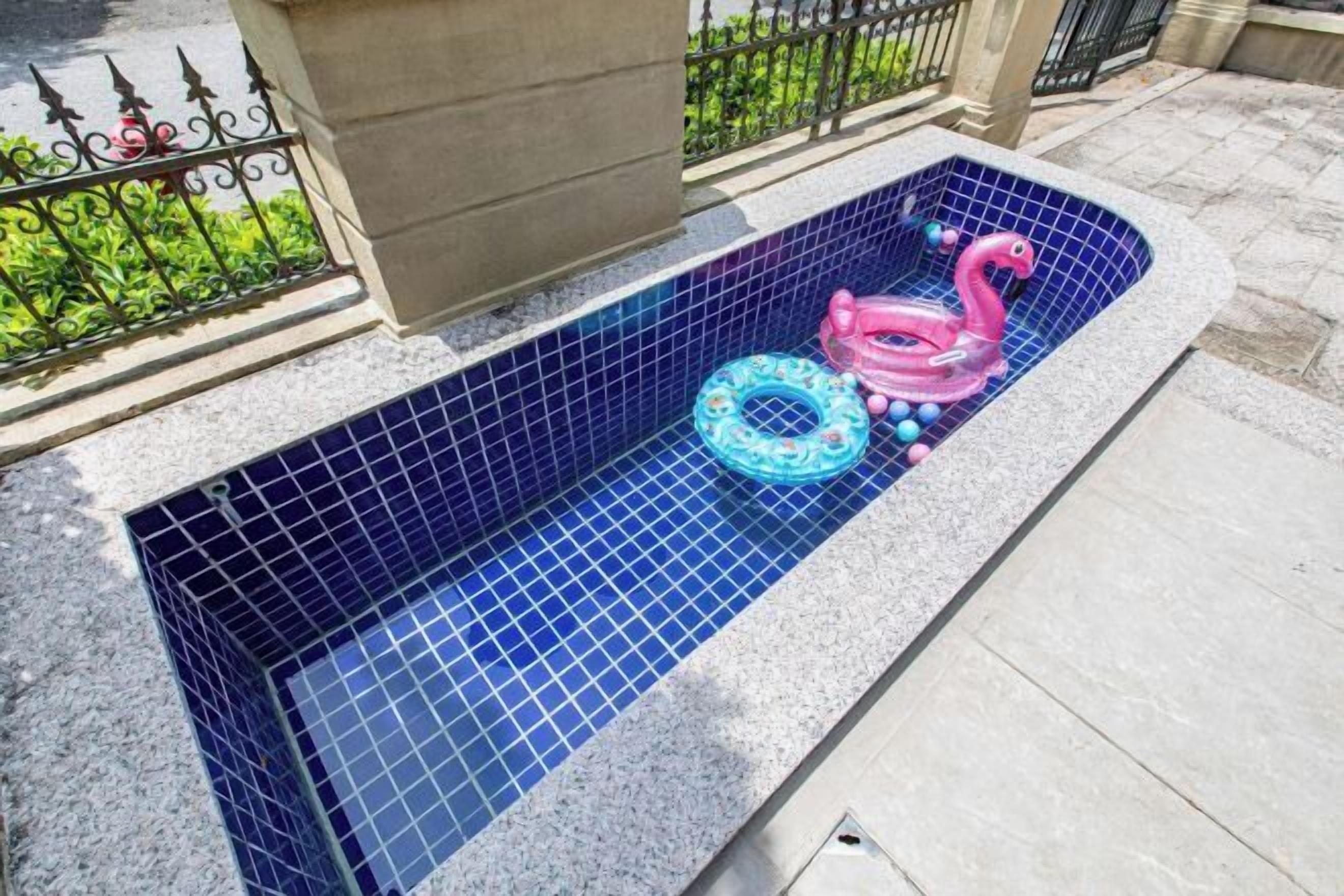 A rectangular outdoor hot tub with blue mosaic tiles, a pink flamingo float, and a blue swirl float.