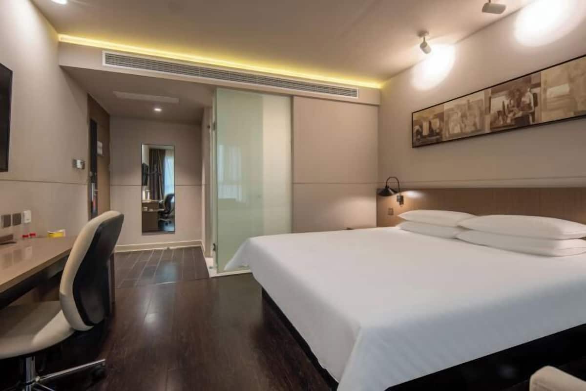 Quarto de hotel com cama, escrivaninha, cadeira e porta de vidro que dá para um banheiro.