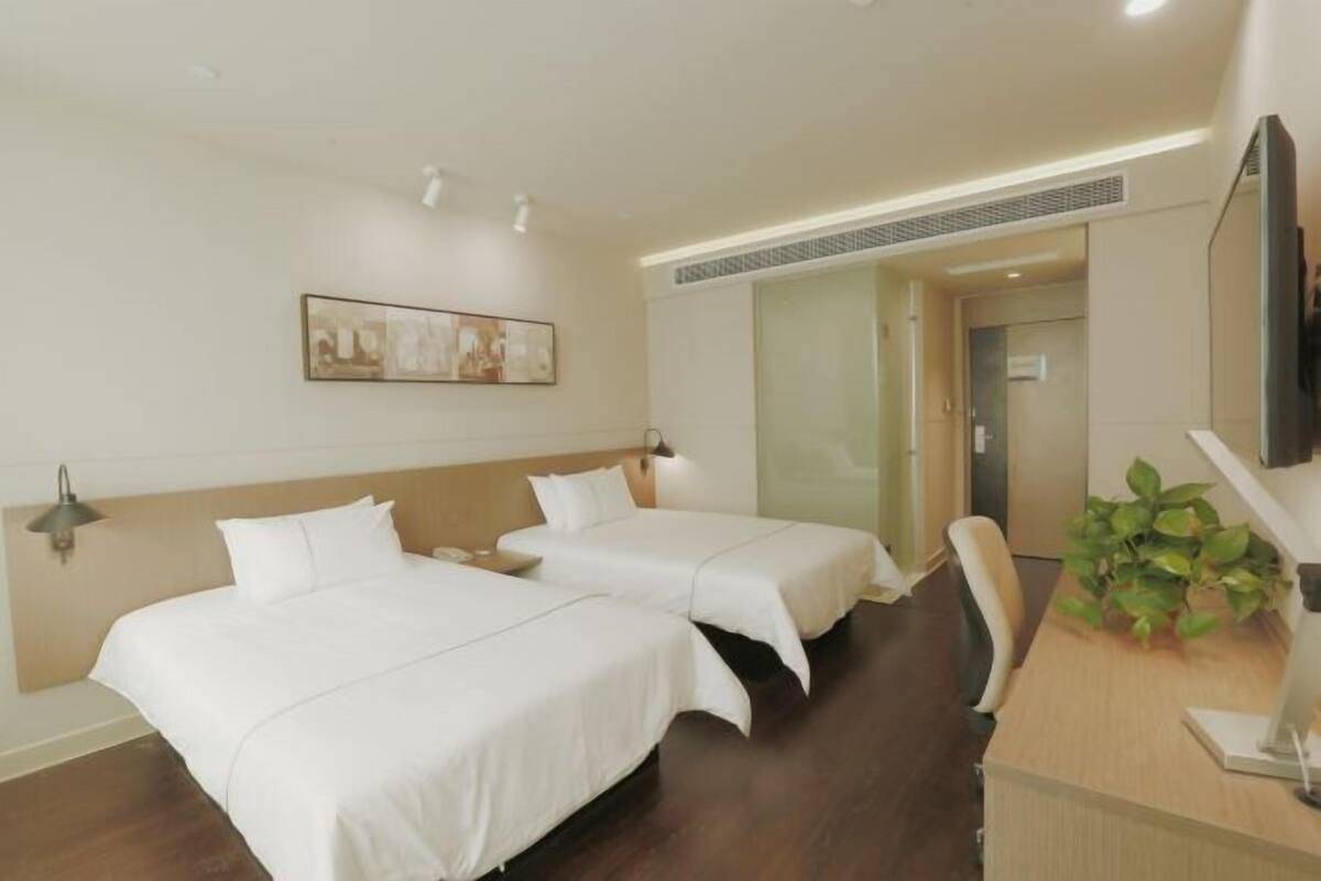 Quarto de hotel com duas camas, uma escrivaninha, uma cadeira, uma televisão e uma planta.