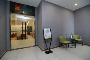 Zona de recepción de un hotel con puerta de cristal, mostrador de recepción y dos sillas verdes.