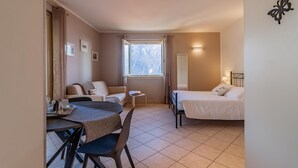 A compact hotel room with a bed, a small desk with a chair, a sofa, a window with a view, and a wall-mounted light fixture. - Incanto Apartment by Wonderful Italy (Bellagio)