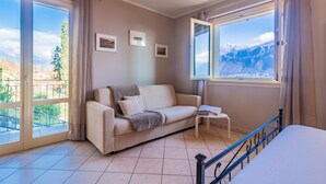 A room with a sofa, a small table, and a bed. There are two windows with curtains, one of which offers a view of mountains. - Incanto Apartment by Wonderful Italy (Bellagio)