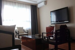 A hotel room with a flat-screen TV mounted on the wall, a wooden coffee table, two armchairs, a sofa, and a large window with curtains. - Jinchang Hotel (Jinchang)