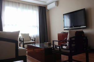 Habitación de hotel con televisor de pantalla plana montado en la pared, una mesa de centro de madera, dos sillones, un sofá y un ventanal con cortinas.