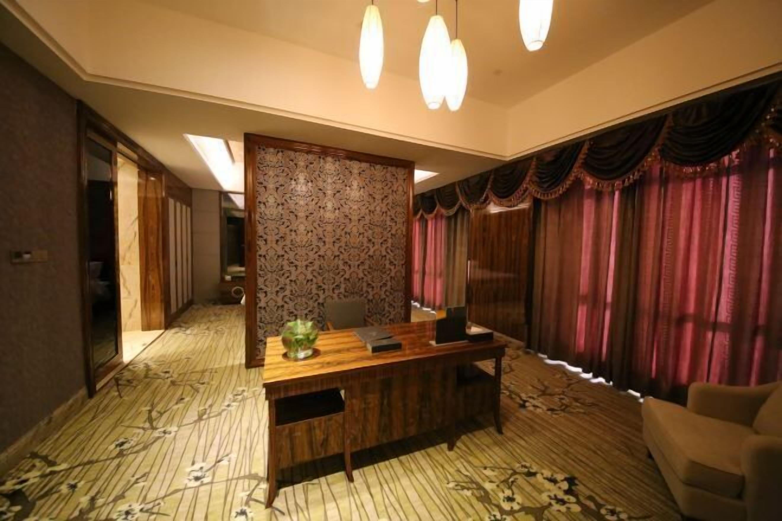 A hotel room with a wooden desk, a chair, a sofa, a patterned carpet, and a large window with curtains.