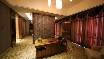 A hotel room with a wooden desk, a chair, a sofa, a patterned carpet, and a large window with curtains.
