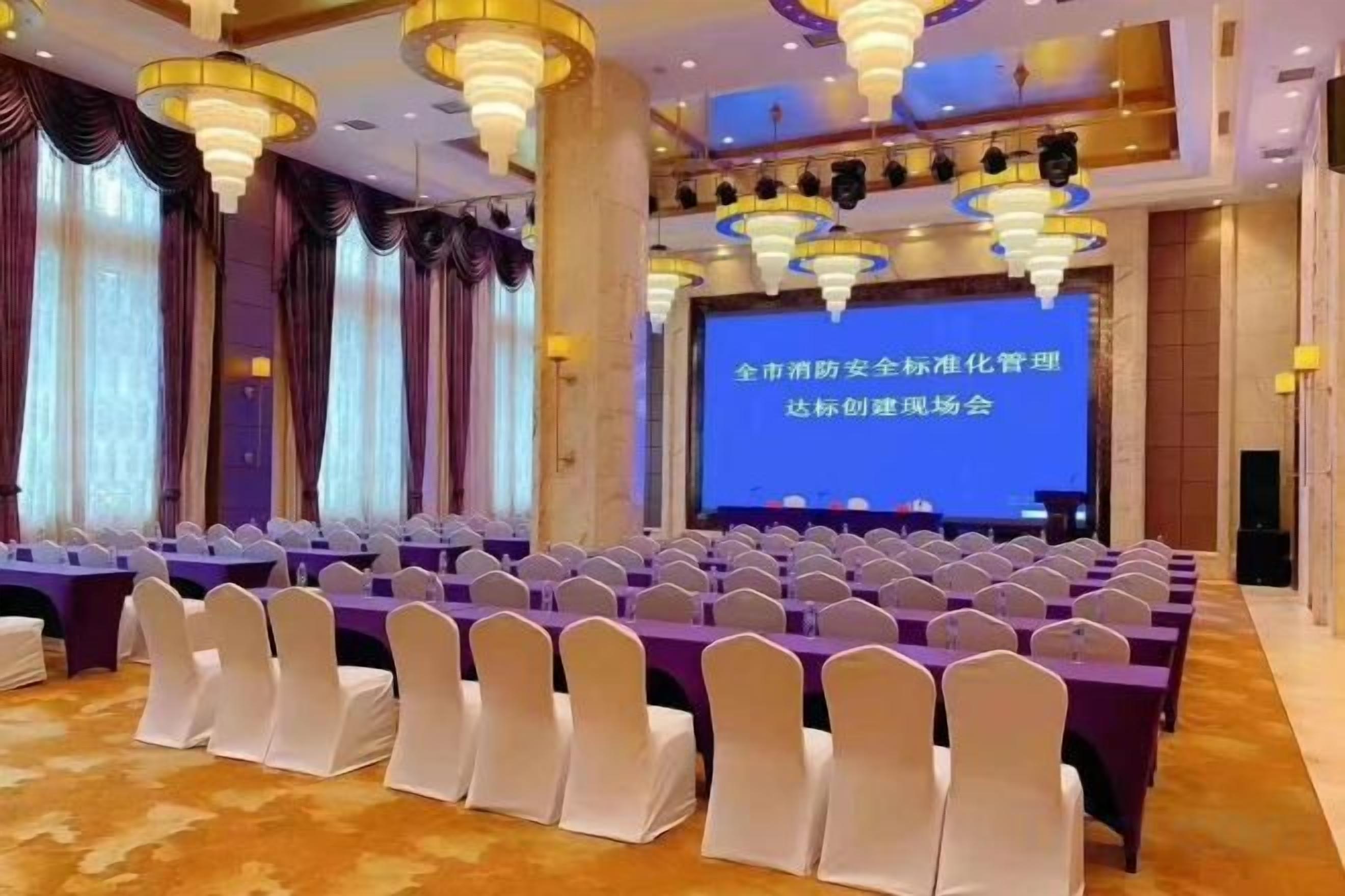 A conference room with rows of white chairs, a stage with a presentation screen, and a chandelier.