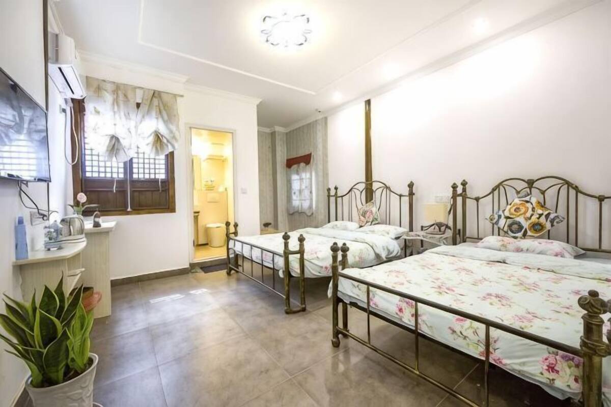 Un dormitorio con dos camas de metal, una mesita de noche, una planta en maceta y una ventana con cortinas.