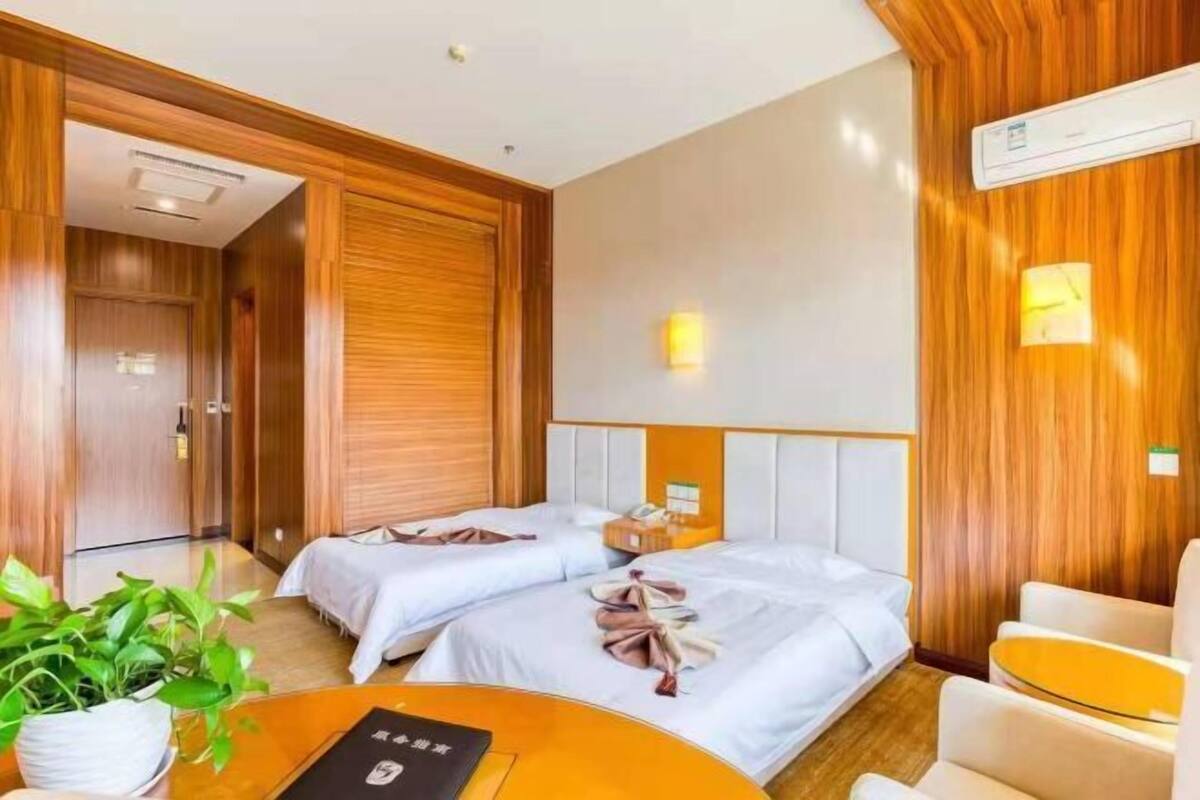 A hotel room with two beds, a small table, a chair, a potted plant, and a wall-mounted air conditioning unit.