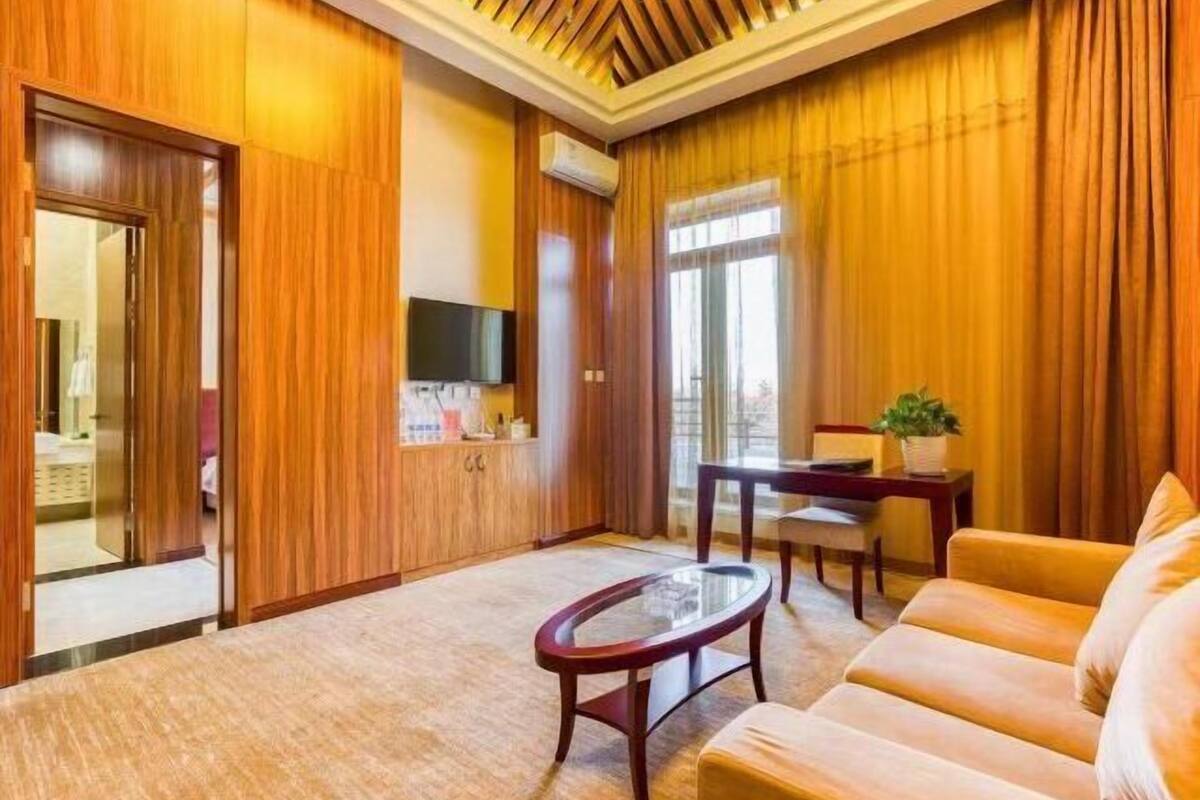 A hotel room with a wooden panel wall, a flat-screen TV mounted above a wooden cabinet, a glass coffee table, a sofa, and a dining table with chairs.