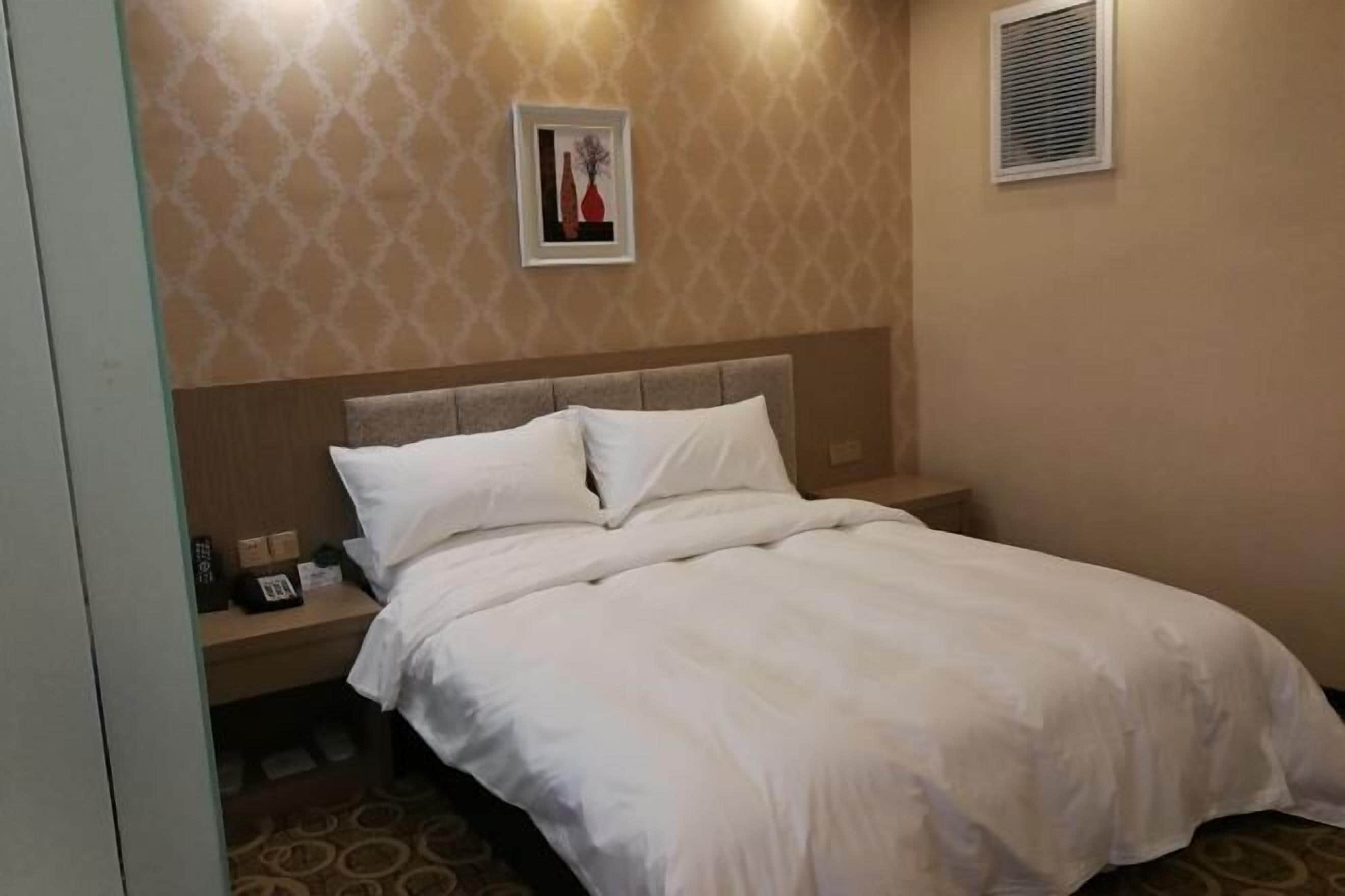 A hotel room with a bed, a nightstand, a framed picture on the wall, and a window with blinds.