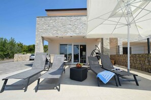 Un espace de détente extérieur moderne, avec des chaises longues, une table ronde ornée de fruits et un parasol blanc.