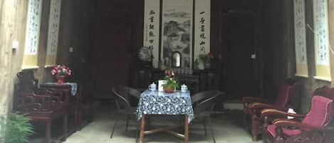 A traditional room with a wooden ceiling, a table with a patterned tablecloth, and chairs. There are calligraphy scrolls on the walls and a decorative hanging in the center.