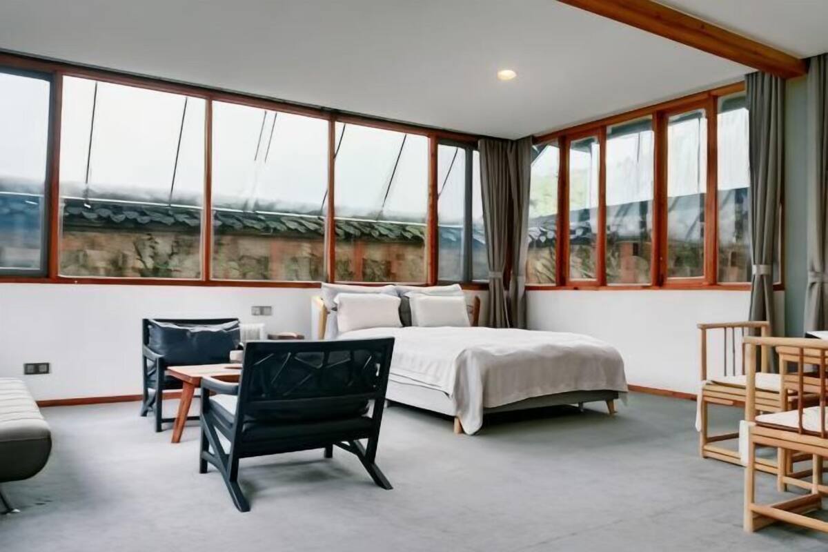 A hotel room with a bed, two chairs, a small table, and large windows.