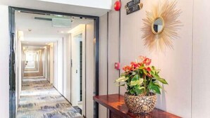 A hotel hallway with a patterned carpet, a wooden reception desk with a potted plant, and a wall-mounted mirror. - Yili Hotel (Kaiping)