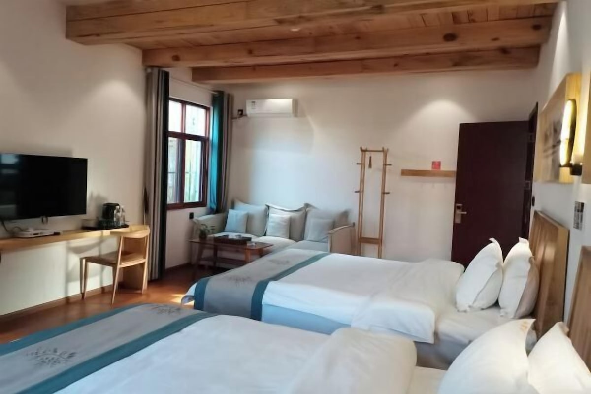 A hotel room with two beds, a desk with a TV, a chair, a window, and a wooden ceiling.