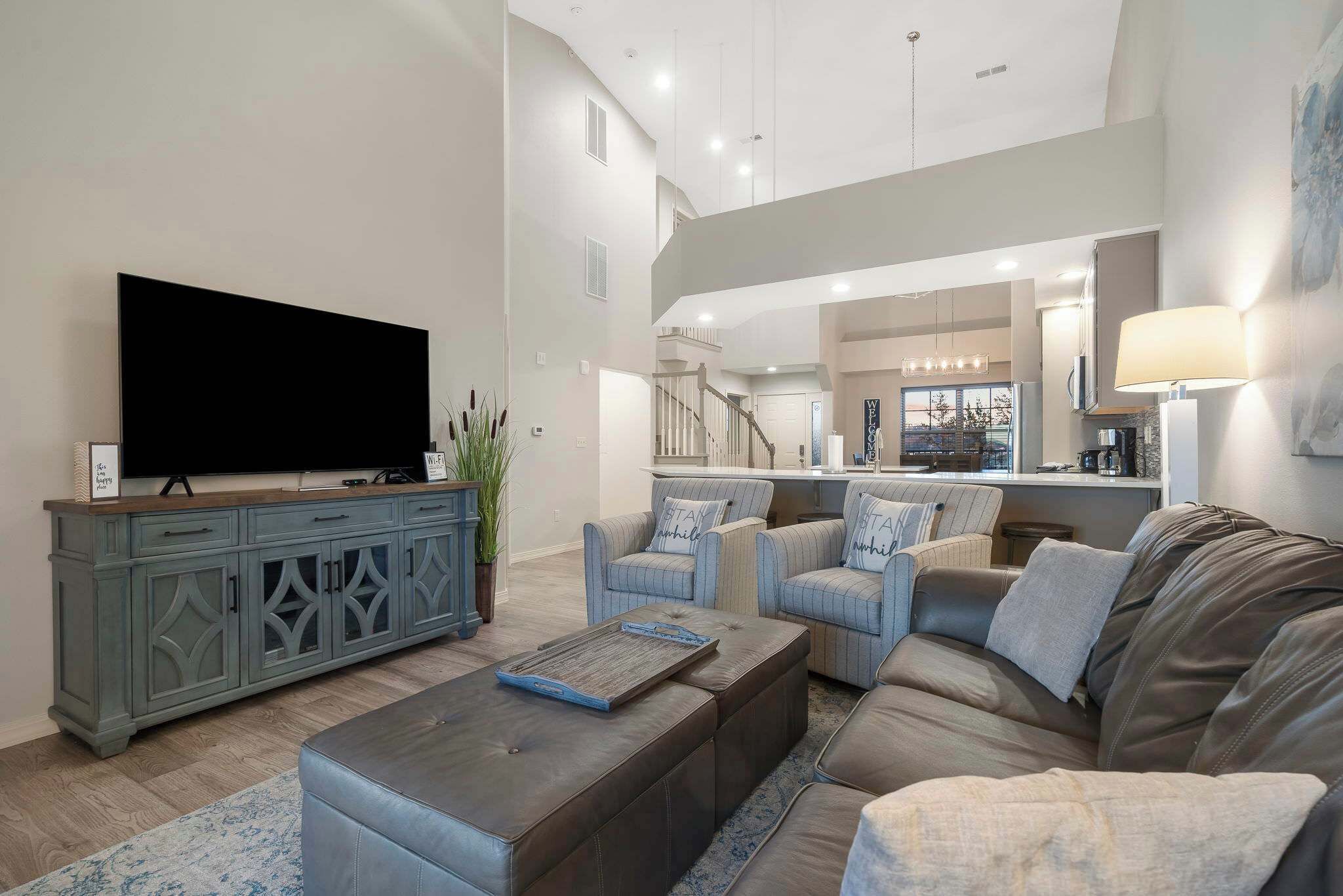 A modern living room with a large flat-screen TV, a grey cabinet, a brown ottoman, and a grey sofa.