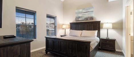 A bedroom with a dark wooden bed, two bedside tables with lamps, a ceiling fan, and two windows with blinds.