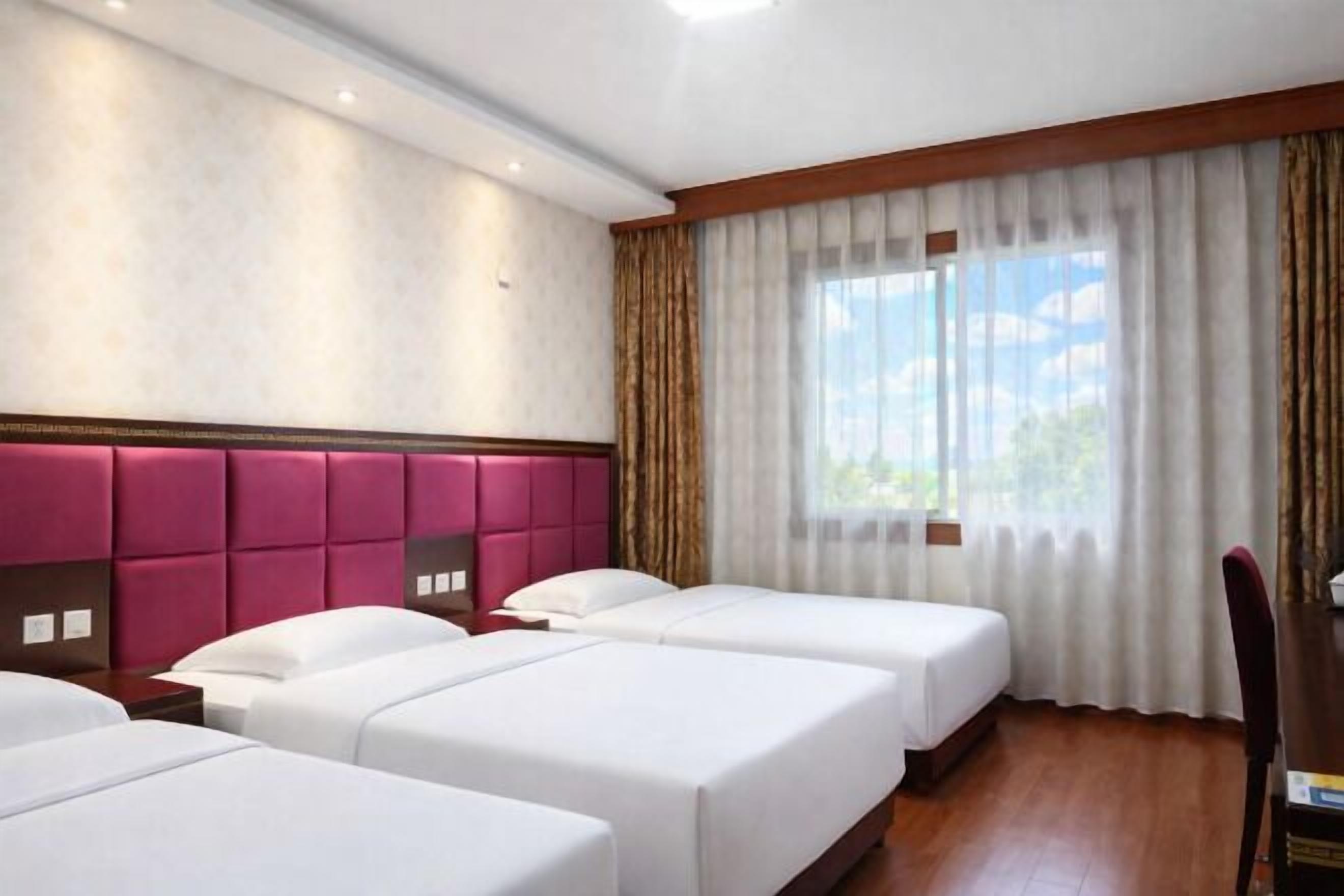 Habitación de hotel con tres camas, un ventanal con cortinas, suelo de madera y cabecera con panel rosa.