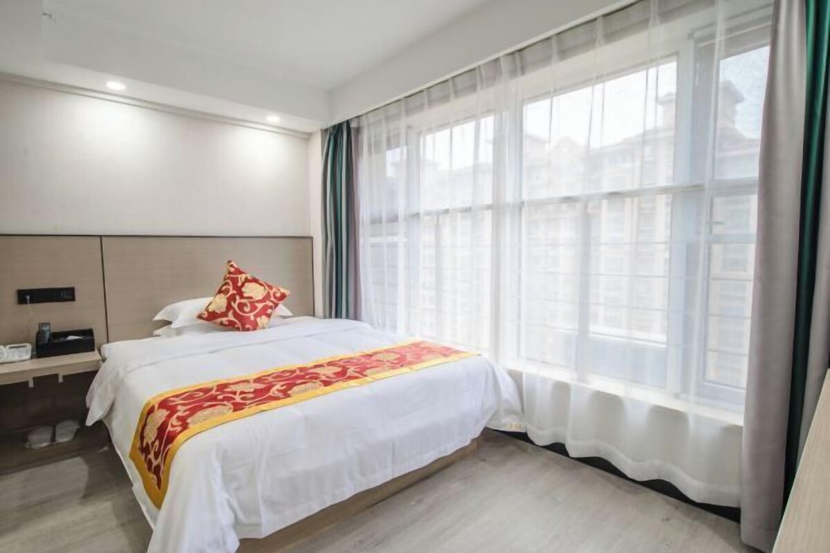 Ein Hotelzimmer mit einem großen Bett, einem Nachttisch, einem kleinen Tisch daneben und einem Fenster mit durchsichtigen Vorhängen.