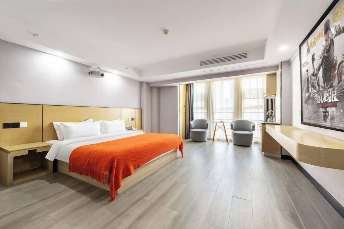 Une chambre d’hôtel avec un lit, une table de chevet, deux fauteuils, un bureau, une grande fenêtre et une affiche au mur.