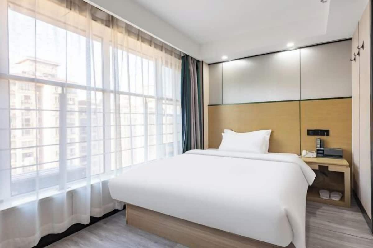 Une chambre d’hôtel avec un grand lit, une table de chevet avec une lampe, une armoire et une fenêtre avec des rideaux légers.