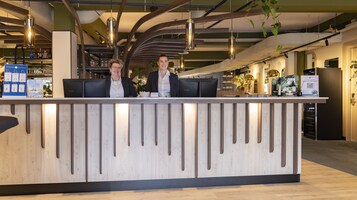 Una recepción con dos empleados, un interior moderno con vigas a la vista y un bar visible.