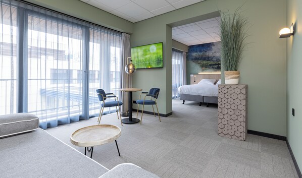 Habitación de hotel con cama, dos sillones, una mesita, televisor y un ventanal con cortinas.