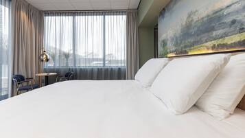 Ein großes Bett mit weißer Bettwäsche, Blick auf die Stadt durch leichte Vorhänge und ein Gemälde an der Wand.