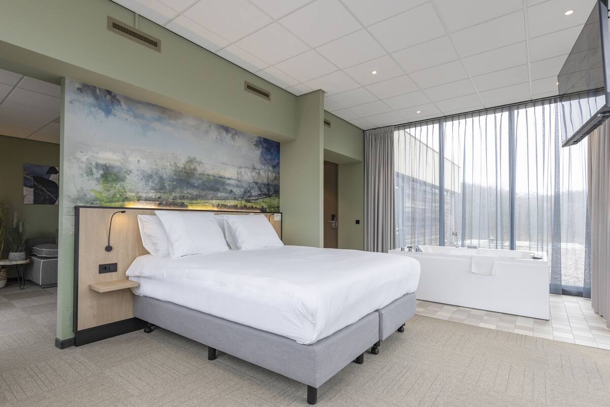 A modern hotel room with a large bed, a separate bathroom with a bathtub, and a city view through large windows.