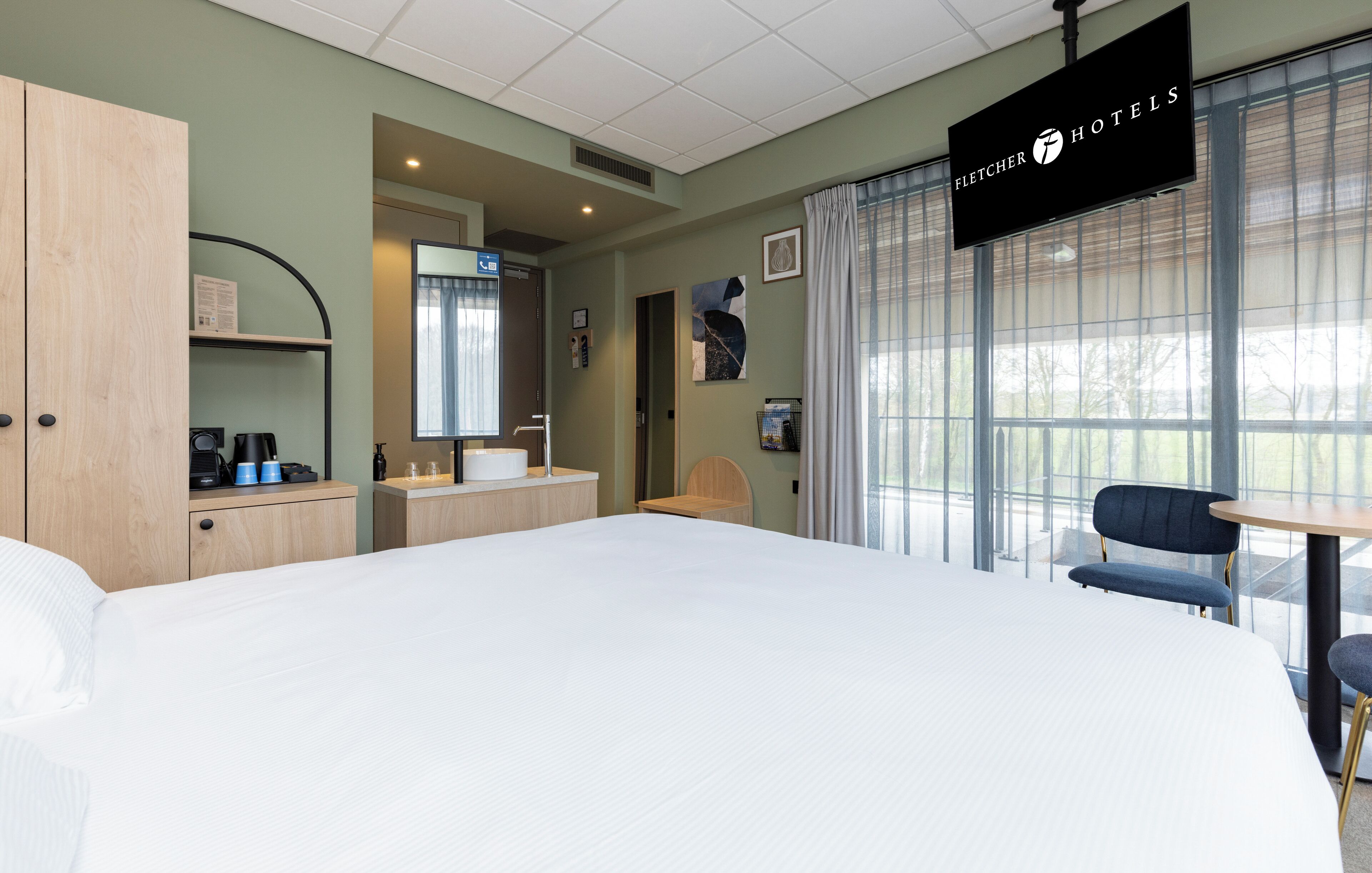 A hotel room with a large bed, a desk with a chair, a small table, and a window with curtains.