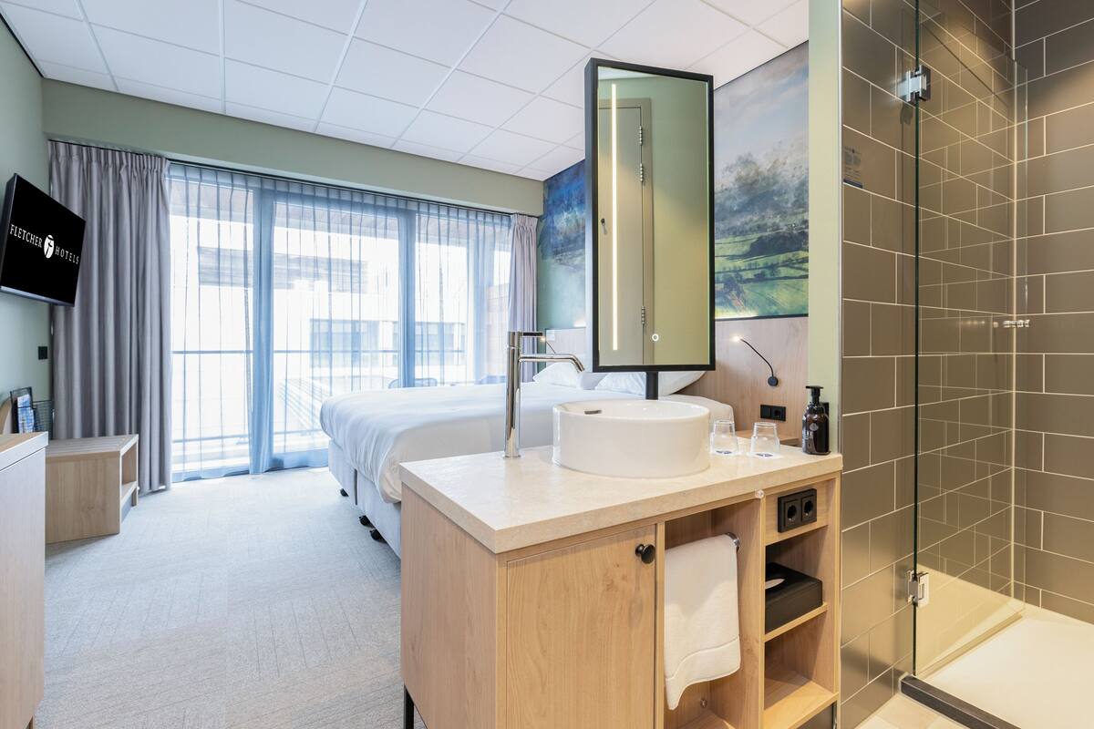 A hotel room with a bed, a wooden vanity with a sink, a large mirror, and a shower area.