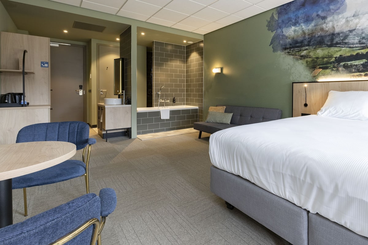 A hotel room with a large bed, a small sofa, a desk with a chair, a bathroom with a shower, and a painting on the wall.